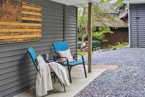 A covered outdoor seating area with two chairs and a table, a wooden wall with a decorative panel, and a gravel path leading to a house. - Lakefront, 5 Min to Colgate, Sleeps 8 (Hamilton)
