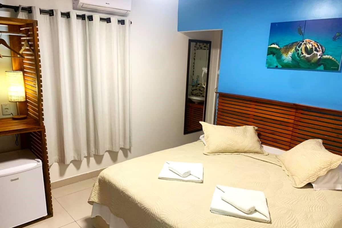 A hotel room with a bed, pillows, a nightstand, a lamp, a window with curtains, and a painting of a sea turtle.