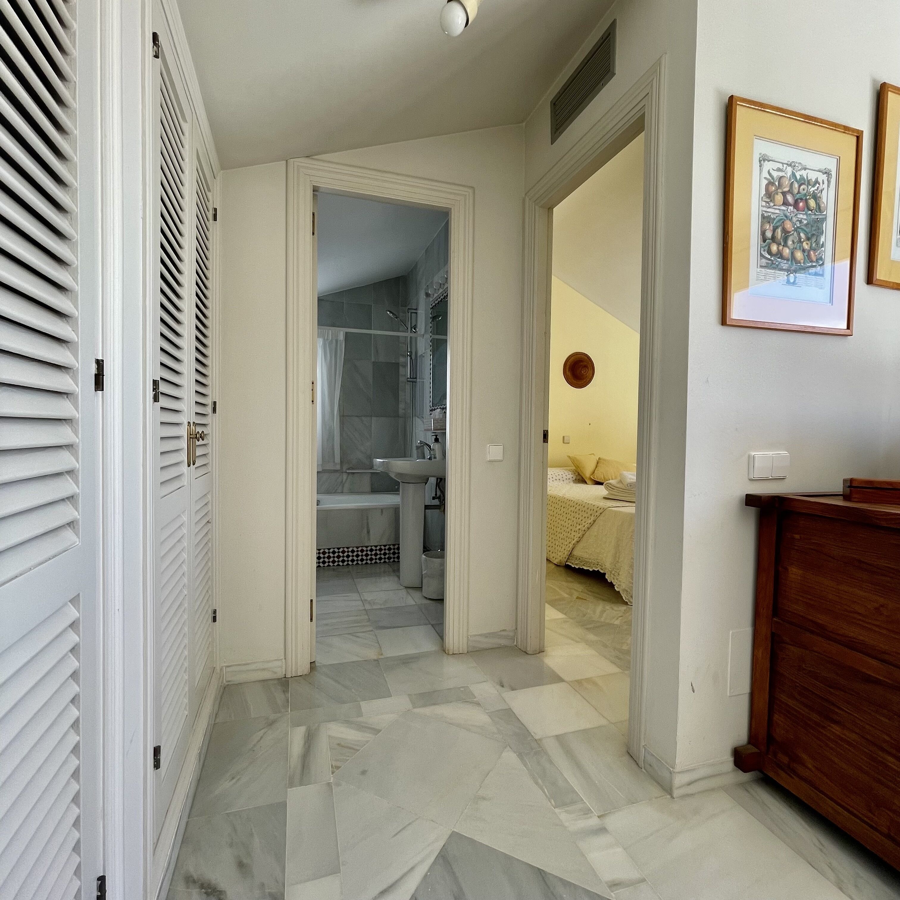 A hallway leading to a bathroom with a shower, a sink, and a bed with a nightstand.