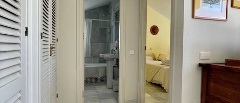 A hallway leading to a bathroom with a shower, a sink, and a bed with a nightstand.