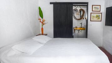 Un dormitorio con cama, mesita de noche, planta, espejo y baño visible a través de una puerta abierta.