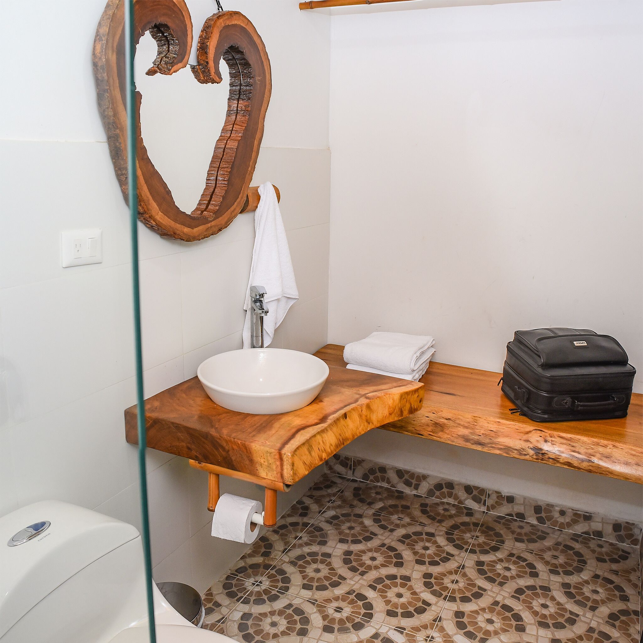 A bathroom with a wooden vanity, a white sink, a towel, a black suitcase, and a toilet.