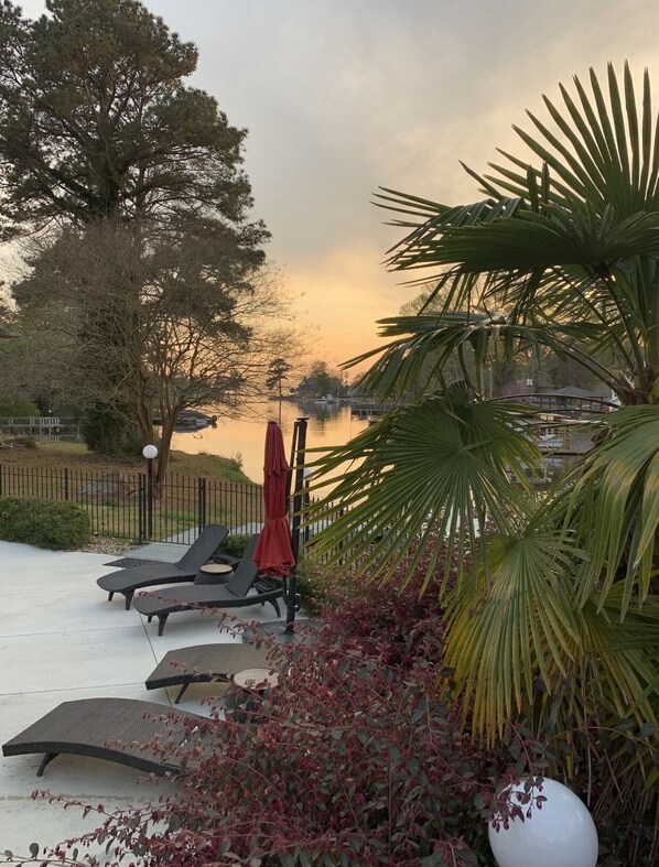 A serene outdoor seating area with lounge chairs and a red umbrella, overlooking a calm body of water and a distant building. - Lake venue w pool, perfect for small venues, weddings, cooperate, business etc. (Irmo)