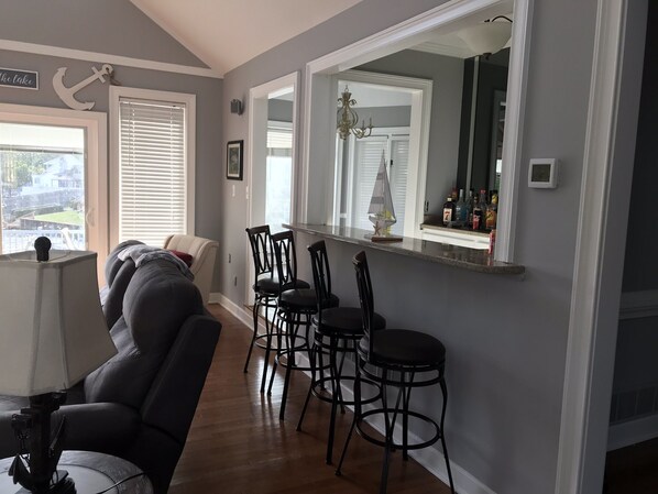 A living room with a sofa, a dining area with bar stools, a kitchen counter with bottles, and a large mirror reflecting the kitchen.
