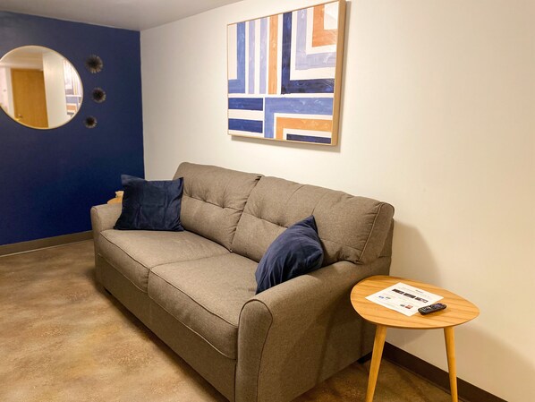A room with a beige sofa, two dark blue pillows, a small wooden side table with a remote control, and a wall with abstract art.