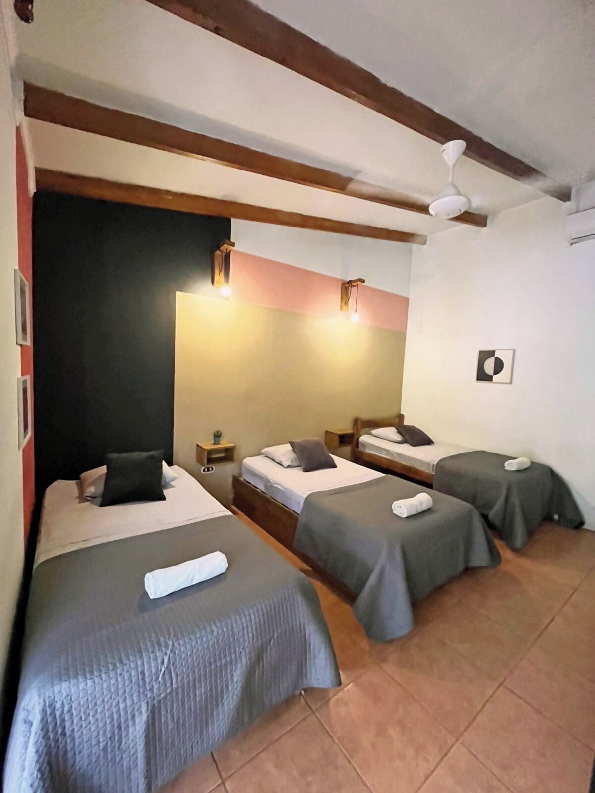 A hotel room with three single beds, each with a pillow and a towel. There are bedside tables with lamps, a ceiling fan, and a framed picture on the wall.
