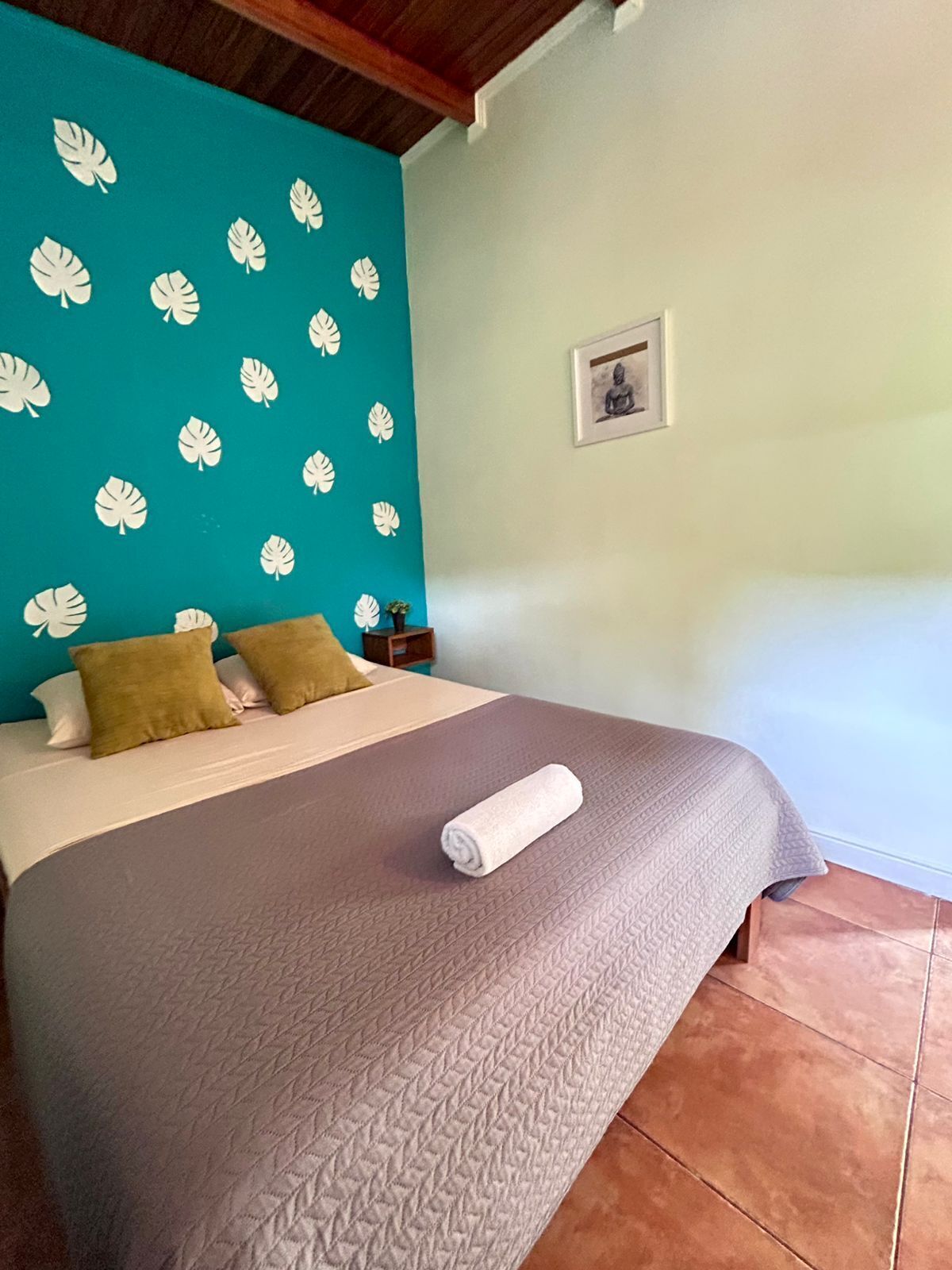 Una cama con pared color turquesa y motivos de hojas blancas, una toalla enrollada y una pequeña estatua de Buda en la mesita de noche.