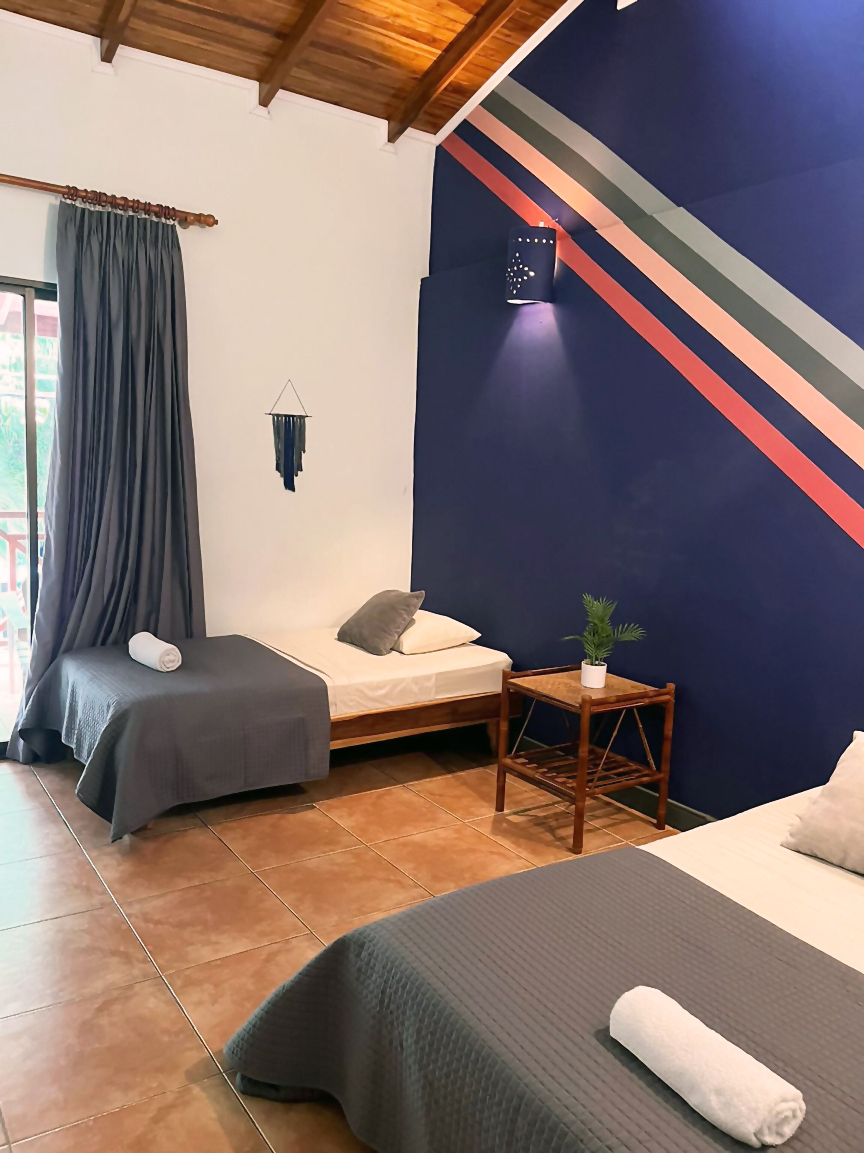 A room with two beds, a small table with a plant, and a wall with a colorful stripe design.