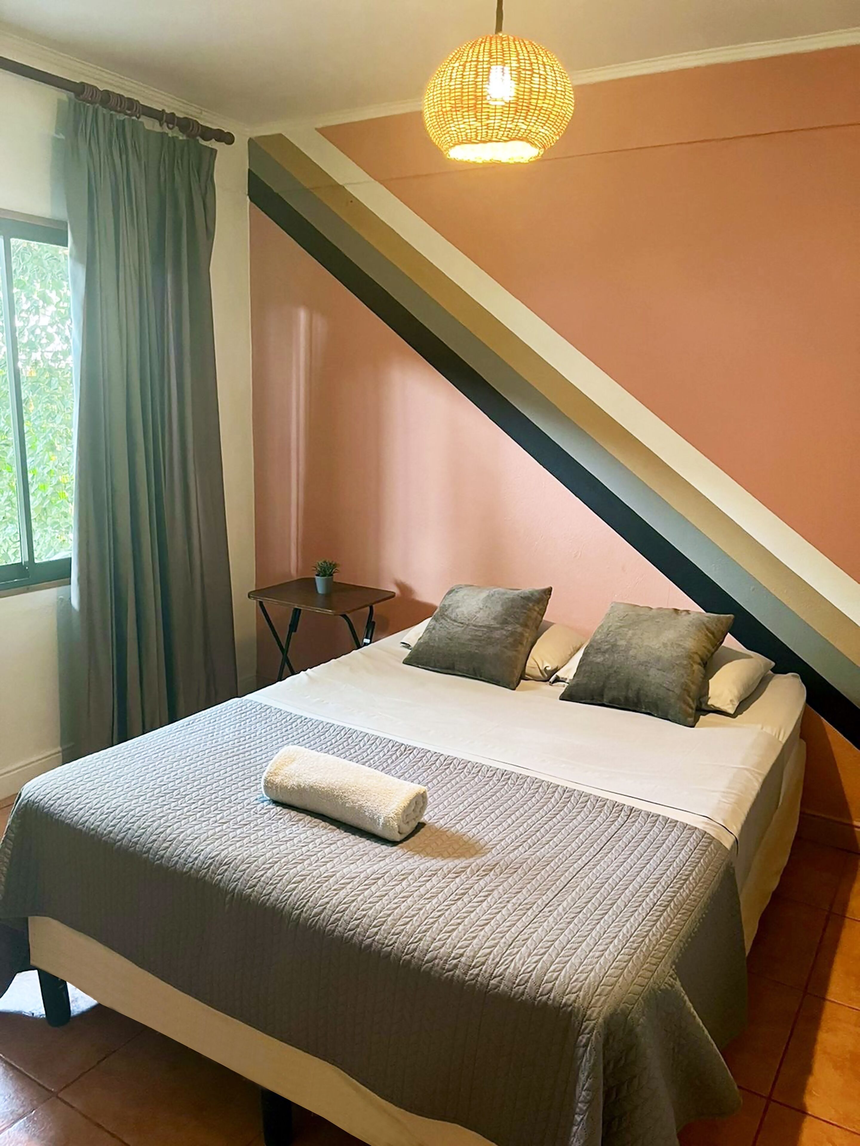 Un dormitorio con cama, almohadas, una mesita de noche, una lámpara colgante y una ventana con cortinas.
