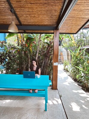 A person sitting at a turquoise bench with a laptop, under a wooden structure with a hanging light, surrounded by lush greenery and a concrete path.