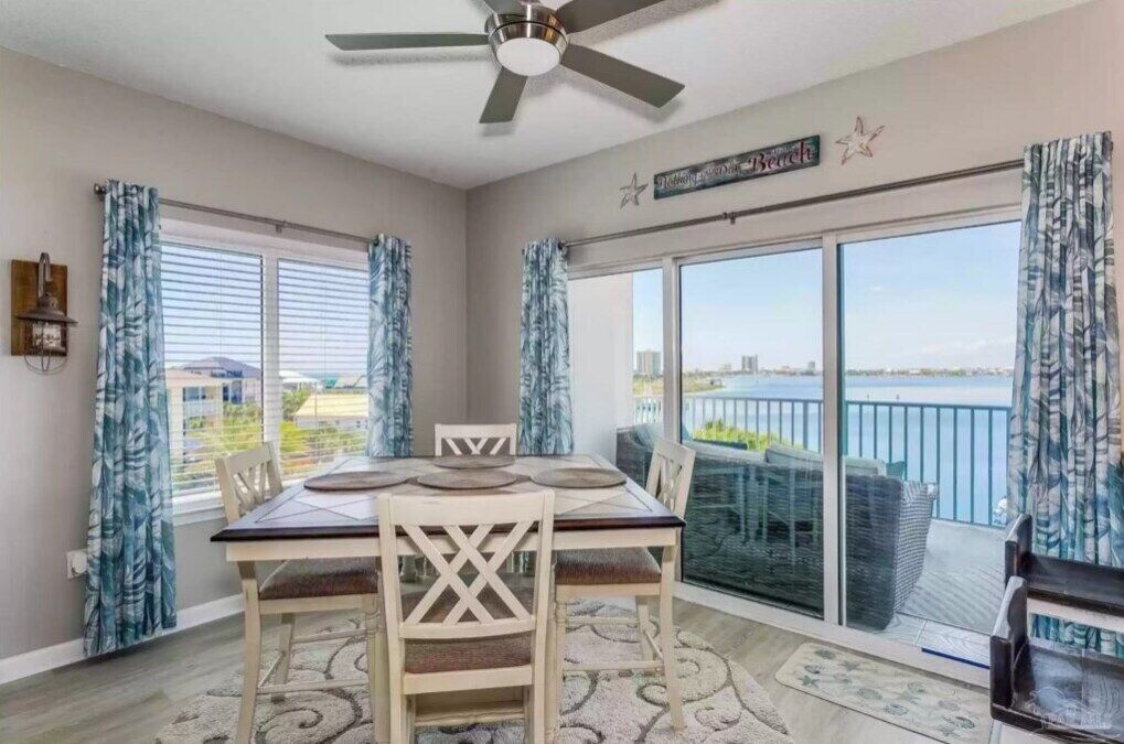 A dining room with a table set for four, a ceiling fan, and a view of the water through large windows.