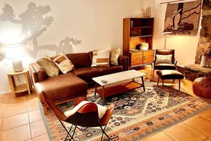 Uma sala de estar com um sofá de couro marrom, um tapete estampado, uma mesa de centro de madeira, uma mesinha lateral, uma estante e uma poltrona.