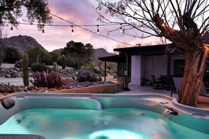 Un bain à remous entouré de plantes désertiques, d’un arbre et d’un bâtiment doté d’un espace extérieur aménagé pour s’asseoir.