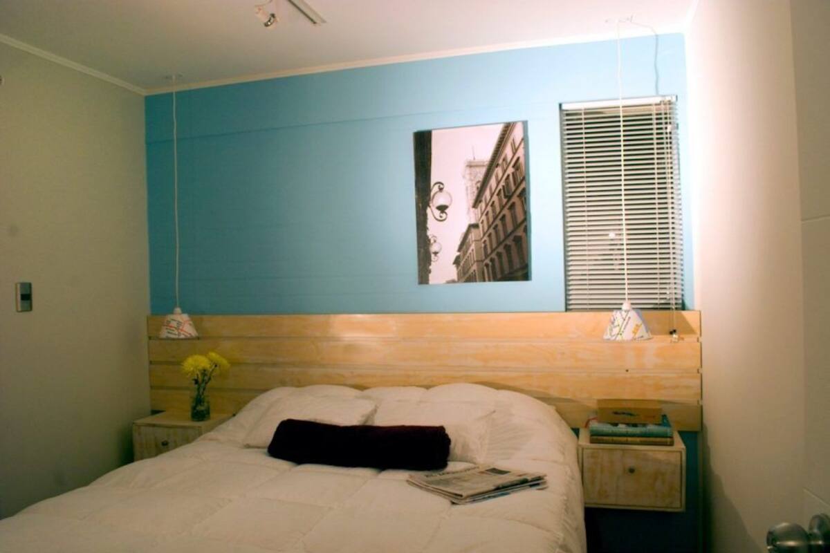 Ein Schlafzimmer mit einem Bett, einem Nachttisch, einem Wandbild und einem Fenster mit Jalousien.