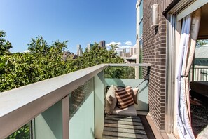 En balkong med ett glasräcke, en bekväm sittgrupp med kuddar och utsikt över en stadssiluett med byggnader och träd.