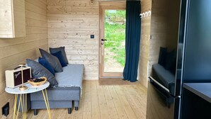 Une pièce de taille modeste aux murs en bois, avec un canapé gris agrémenté de coussins, une petite table sur laquelle se trouve un haut-parleur, et un réfrigérateur noir.