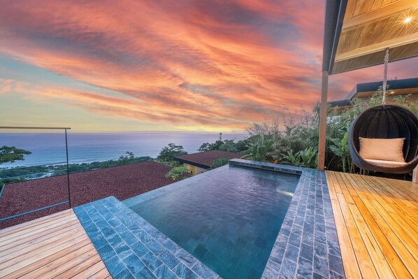 A rooftop infinity pool with a wooden deck and a hanging chair, overlooking a scenic ocean view at sunset.