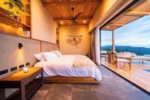 A modern bedroom with a wooden ceiling fan, a bed with white linens, a bedside table with a lamp, and a view of the outdoors through large windows.