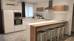 A modern kitchen with white cabinetry, a built-in oven, a coffee maker, and a wooden bar counter with bar stools. - Pretty air-conditioned single-storey villa with swimming pool (Cabestany)