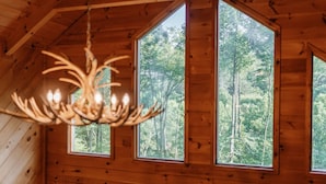 A wooden cabin interior with a chandelier and three windows offering a forest view. - Pet-Friendly Blue Ridge Cabin on 9 Acres! Views, Theater, Game Room, Trails! (Blue Ridge)