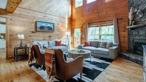 A cozy cabin interior with wooden walls, a stone fireplace, and a large window overlooking greenery. - Pet-Friendly Blue Ridge Cabin on 9 Acres! Views, Theater, Game Room, Trails! (Blue Ridge)