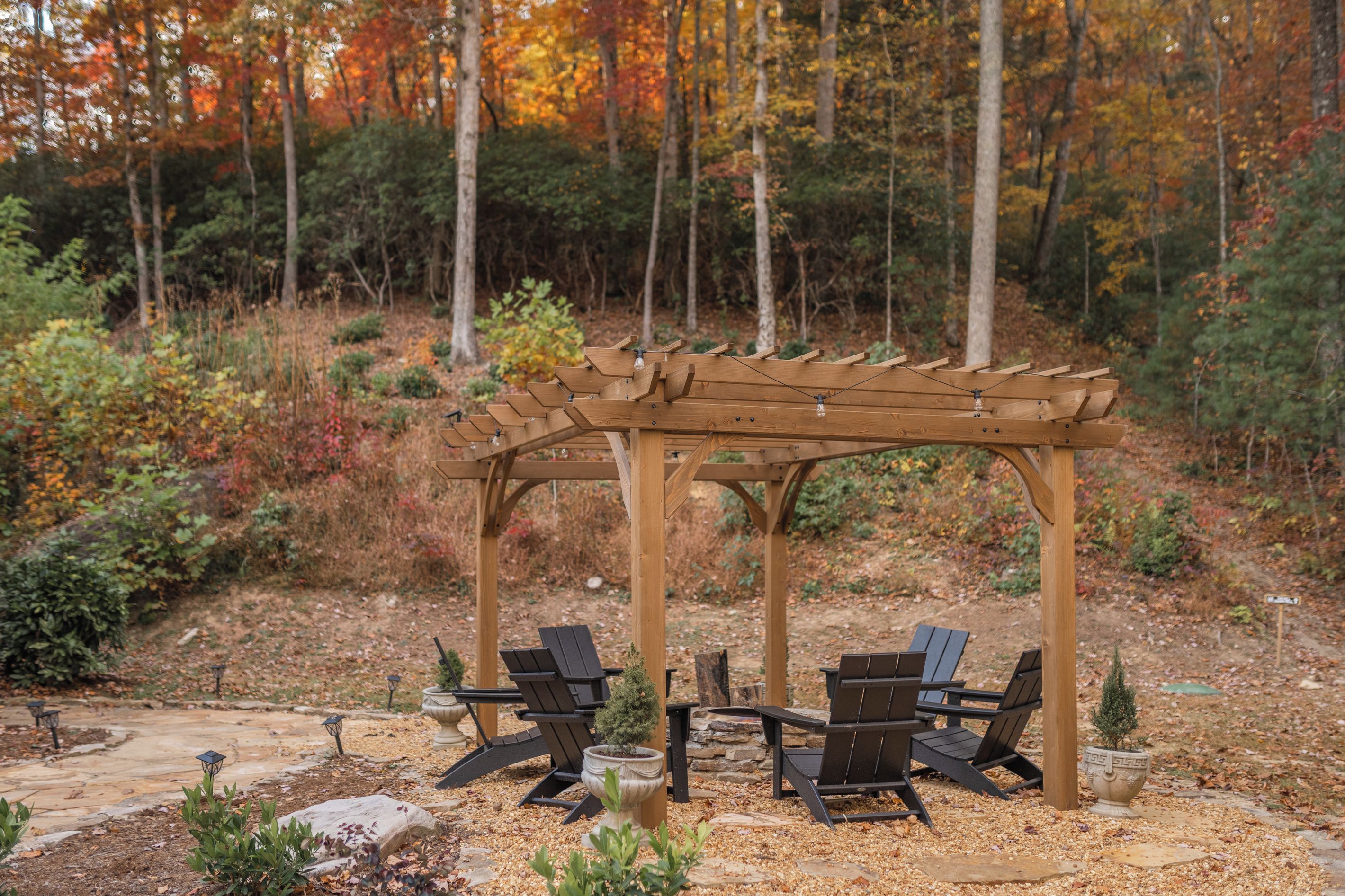 A wooden pergola with four chairs and a table, surrounded by a garden with autumn foliage.
