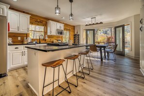 A modern kitchen with a large island, bar stools, and a view of autumn foliage through the windows.