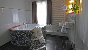A bathroom with a large bathtub, a sink, and a mirror. There is a potted plant on the windowsill and a towel on the floor.