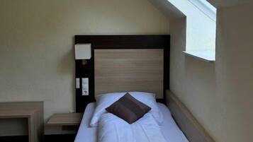 A single bed with a wooden headboard, a bedside table, and a small window providing natural light.