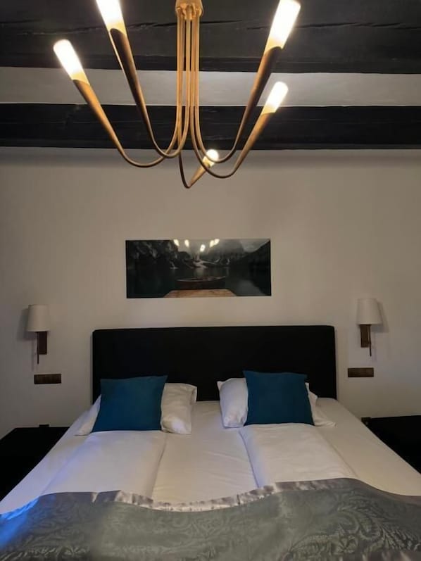 Una cama doble con ropa de cama blanca y azul, un cabecero negro y una araña con múltiples luces.