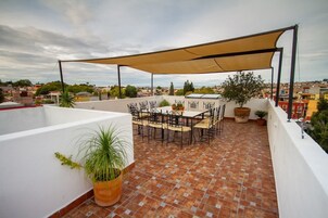 Terraza en la azotea con mesa y sillas, rodeada de plantas en macetas y una gran pérgola.