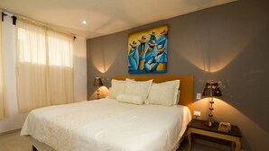 Une chambre bien éclairée, avec un grand lit, deux lampes de chevet, un tableau au mur et une table de chevet sur laquelle se trouve un livre.