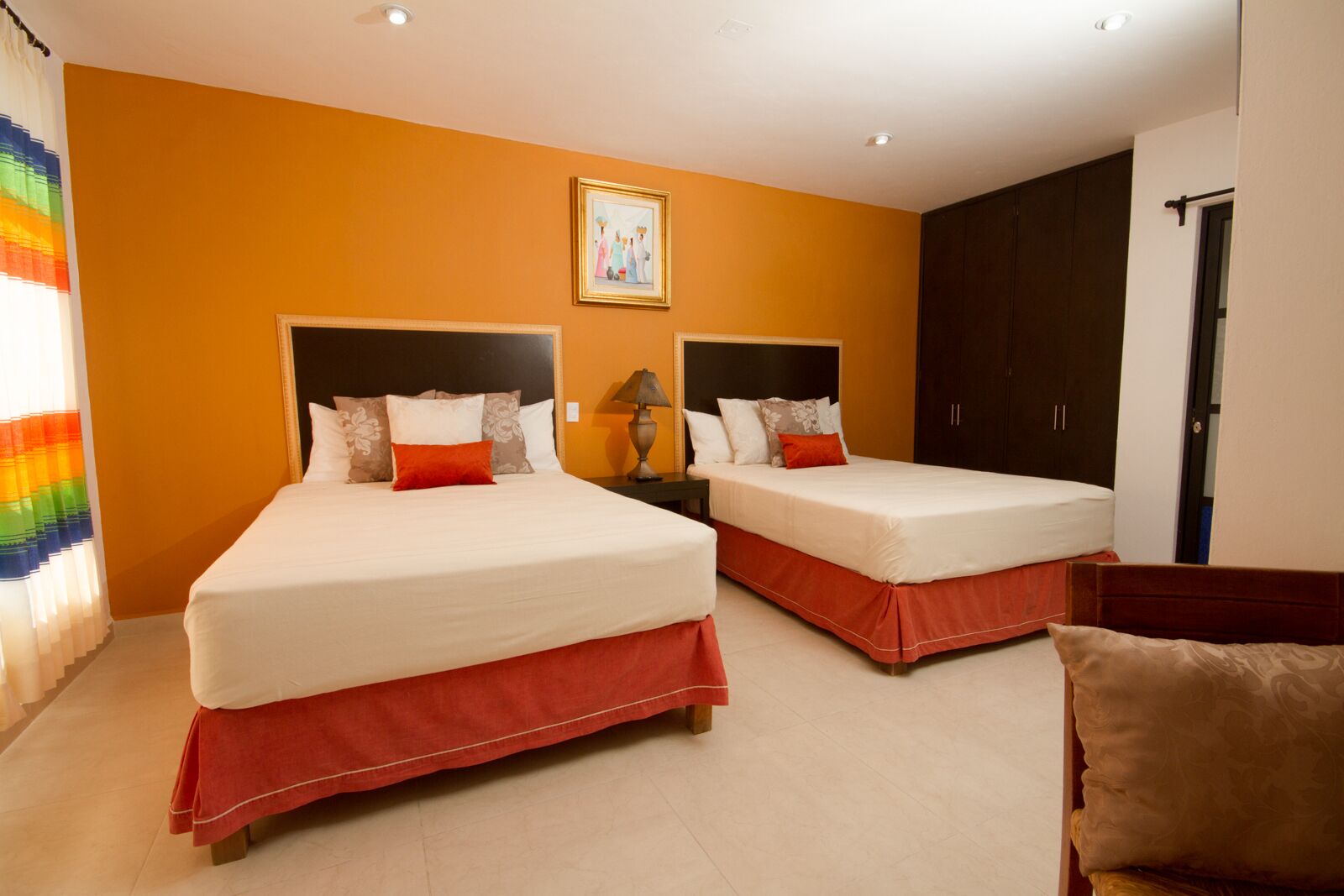 Habitación de hotel con dos camas, cada una con falda roja, ropa blanca y varios almohadones. Entre las camas, una mesita de noche con lámpara. La habitación tiene una pared de acento naranja y un cuadro enmarcado sobre las camas.