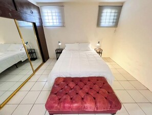 Un dormitorio con un espejo grande, una cama, dos mesitas de noche con lámparas y un otoman tapizado en rojo.