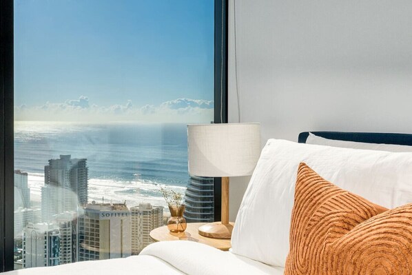 A hotel room with a large window offering a city view, a bed with white and brown pillows, and a bedside table with a lamp. - The Darling of Broadbeach - with spectacular views (Broadbeach)