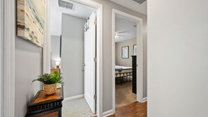 Un couloir avec une plante posée sur une console, une porte donnant sur une pièce où se trouve un lit, et un coin repas avec une table et des chaises.