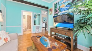 A living room with a teal wall, a wooden coffee table with a fish painting, a television on a wooden stand, and a large plant.