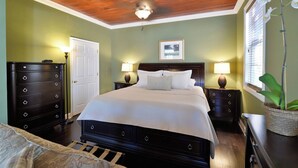 Une chambre à coucher comprenant un lit, deux tables de chevet, une commode, un ventilateur de plafond et un tableau encadré accroché au mur.