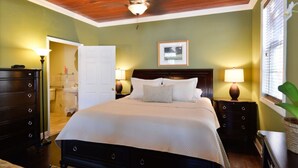 Un dormitorio con techo de madera, una cama con ropa de cama blanca, dos mesitas de noche con lámparas, una mesilla auxiliar, un aparador y una ventana con persianas.