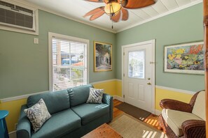 Une pièce avec un canapé vert, un fauteuil en osier, un ventilateur de plafond et une porte avec une fenêtre.