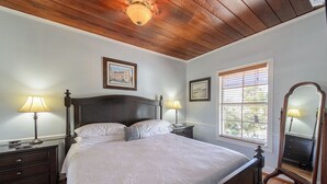 Un dormitorio con techo de madera, una cama grande con ropa blanca, dos mesitas de noche con lámparas y un espejo.