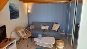 A cozy living room with a grey sofa, a wicker chair, a small white table, and a lamp. - 4-person house in private residence with swimming pool, 600 m from the sea (Bretignolles-sur-Mer)