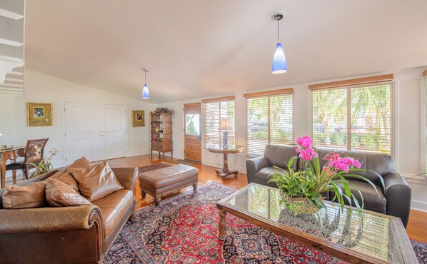 A spacious living room with a leather sofa, ottoman, and a glass coffee table. There is a patterned rug on the floor, a wooden bookshelf, and a lamp on a side table.