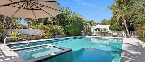 A swimming pool with a sun umbrella, surrounded by lush greenery and a white fence.