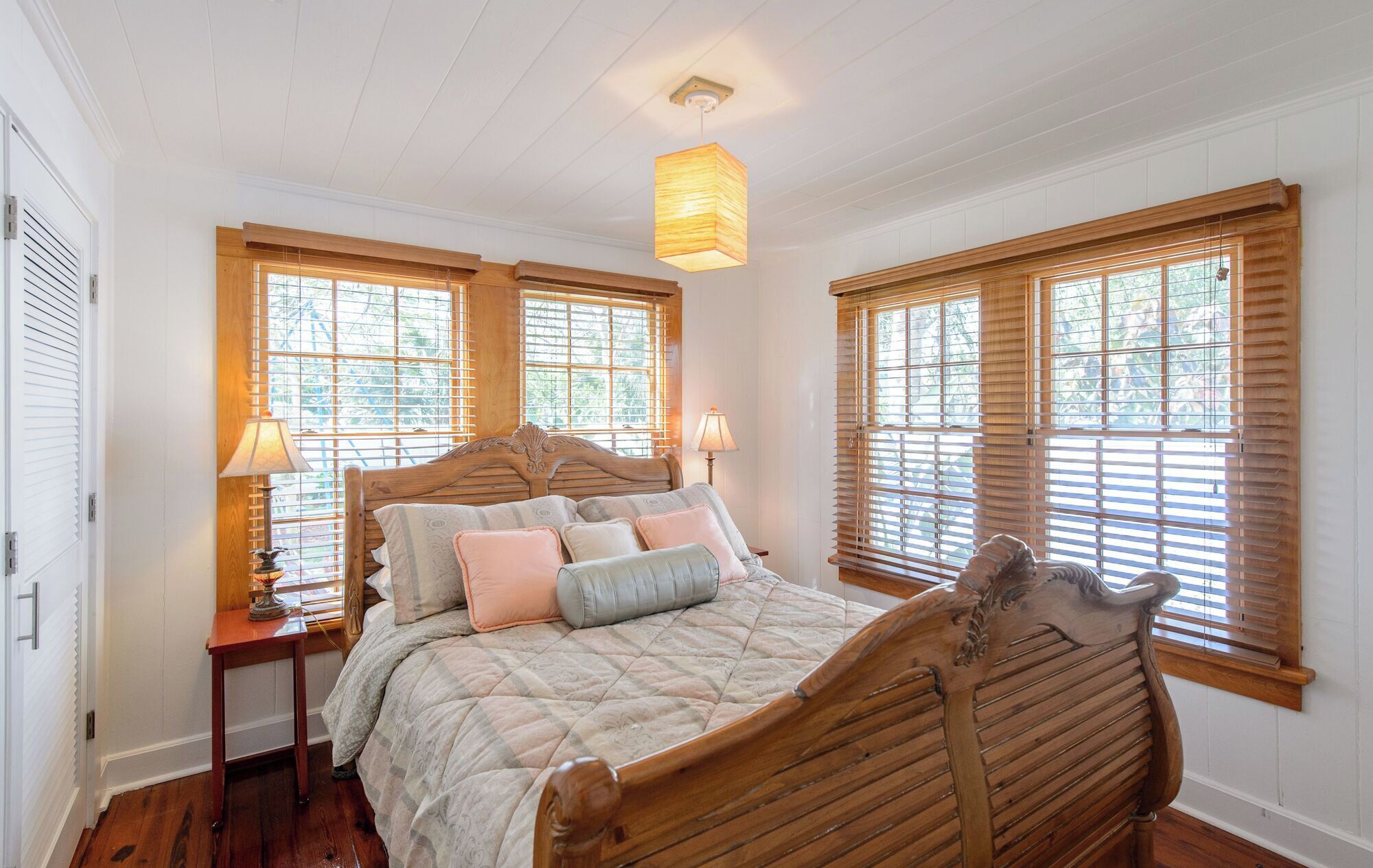 A bedroom with a wooden bed, two bedside tables with lamps, and large windows with blinds.