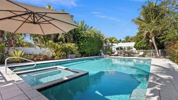 A swimming pool with a sun umbrella, surrounded by lush greenery and a white fence.