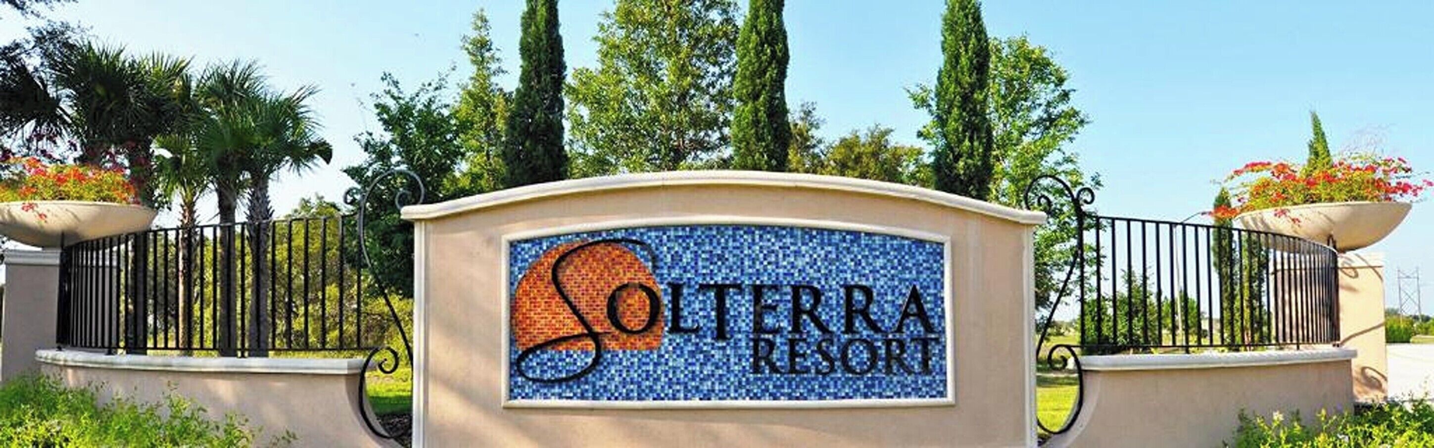 A sign for Solterra Resort with a decorative tile design, surrounded by a black metal fence and potted plants.