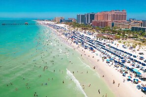 A beach with numerous umbrellas and people, a pier extending into the water, and several high-rise buildings in the background.