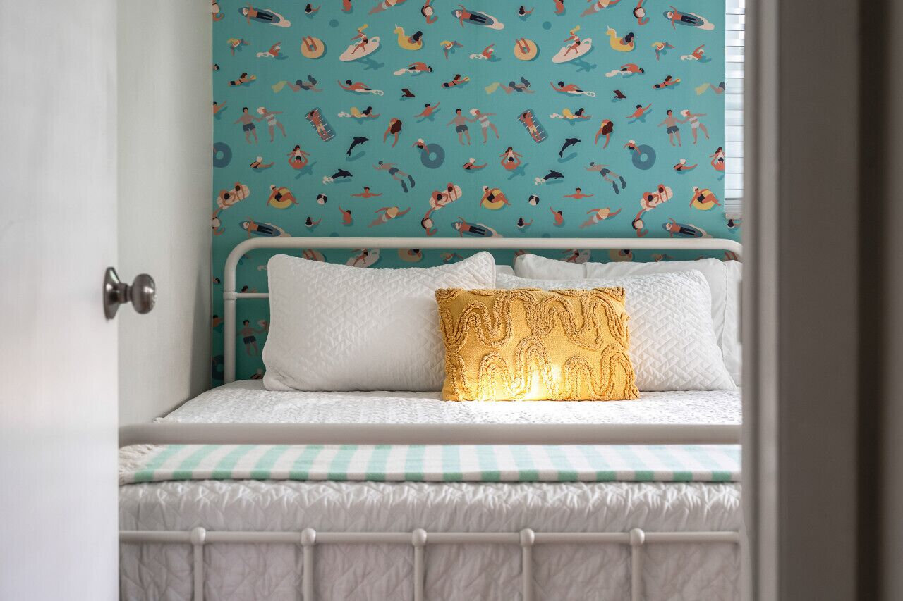 Un lit avec une literie blanche et un oreiller doré, un mur de couleur turquoise orné d’illustrations représentant des personnes pratiquant diverses activités, et une porte blanche.