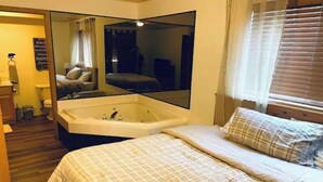 Un dormitorio con un espejo grande, una cama con ropa de cama escocesa, un baño con bañera y una ventana con persianas.
